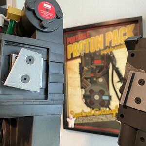 May include: Close-up of a black and gray Ghostbusters proton pack replica with a gray plastic detail on the side. The pack is in front of a framed poster that reads "Portable Proton Pack".
