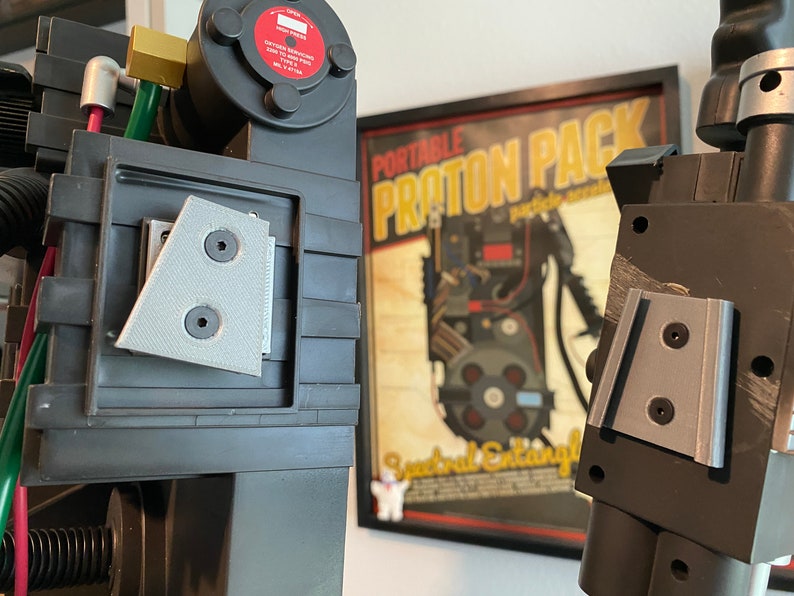 May include: Close-up of a black and gray Ghostbusters proton pack replica with a gray plastic detail on the side. The pack is in front of a framed poster that reads "Portable Proton Pack".