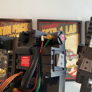 May include: A black and gray Ghostbusters proton pack replica with a warning label, hoses, and wires. The pack is in front of a poster that reads "Futuristic Ecto 1 Lab Headquarters".