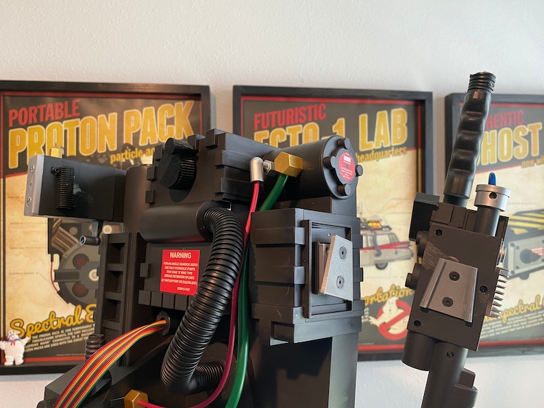 May include: A black and gray Ghostbusters proton pack replica with a warning label, hoses, and wires. The pack is in front of a poster that reads "Futuristic Ecto 1 Lab Headquarters".