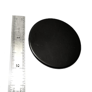 May include: A black, oval-shaped object, approximately 5 cm in diameter, is positioned next to a ruler. The ruler is marked with inch measurements. The object appears to be a lid or a small disc.