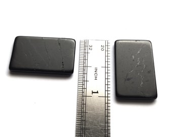 May include: Two black rectangular pieces of polished obsidian, each measuring approximately 1 inch in length, are placed on a white surface next to a metal ruler.