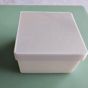 May include: A rectangular white storage box with a textured, slightly shimmering lid. The container features smooth, solid white sides and a flat top surface with a fine, granular finish, presenting a clean and simple design for organising small items.