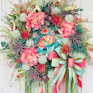 May include: A vibrant, handcrafted wreath featuring a sea turtle design. The wreath is adorned with pink hydrangeas, succulents, and greenery. A large bow with pink, light blue, and green ribbons adds a decorative touch. This wreath is perfect for adding a touch of coastal charm to any home.