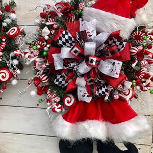 May include: A Christmas wreath with a red Santa hat, white fur trim, and black elf shoes. The wreath is decorated with red and white candy canes, red and white peppermint candies, and a black and white checkered bow.