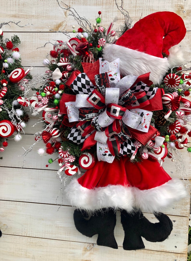 May include: A Christmas wreath with a red Santa hat, white fur trim, and black elf shoes. The wreath is decorated with red and white candy canes, red and white peppermint candies, and a black and white checkered bow.