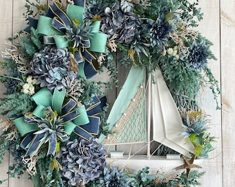 May include: A blue and green wreath with a white sailboat in the center. The wreath is decorated with hydrangeas, succulents, and eucalyptus. There are also two blue and green bows on the wreath.