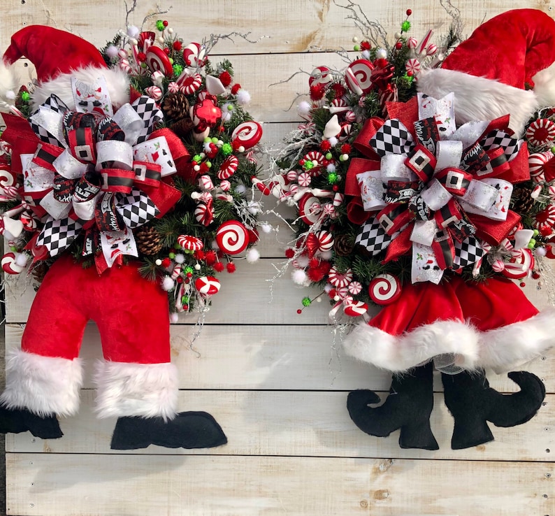 May include: Two Christmas wreaths decorated with red and white candy canes, green and white greenery, and a red and white bow. The wreaths are shaped like Santa Claus with red velvet pants and a white fur trim.