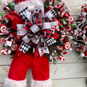 May include: A Christmas wreath featuring a Santa Claus figure with a red hat, red pants, and white fur trim. The wreath is decorated with red and white candy canes, red and white swirl candies, and a large black and white checkered bow.