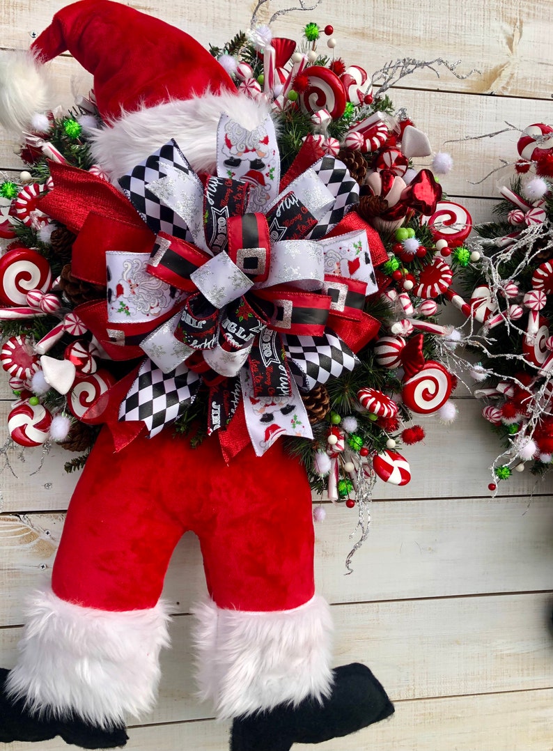 May include: A Christmas wreath featuring a Santa Claus figure with a red hat, red pants, and white fur trim. The wreath is decorated with red and white candy canes, red and white swirl candies, and a large black and white checkered bow.