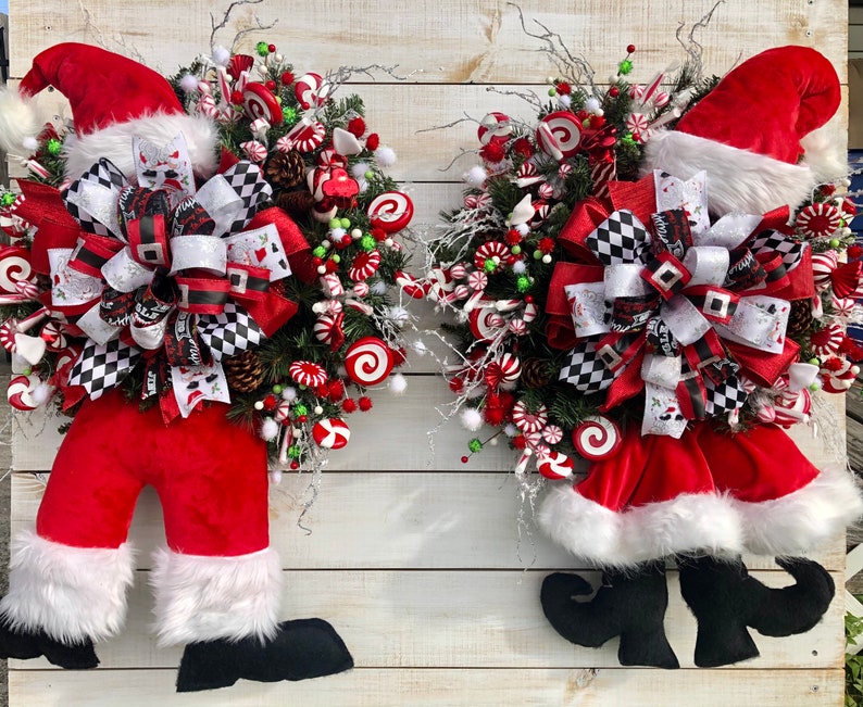 May include: Two Christmas wreaths with Santa hats, red velvet, and white fur trim. Each wreath is decorated with a black and white checkered bow, candy canes, and pine cones.