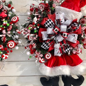 May include: A Christmas wreath decorated with candy canes, red and white swirl candies, and a Santa hat with black boots. The wreath is adorned with a black and white checkered bow.