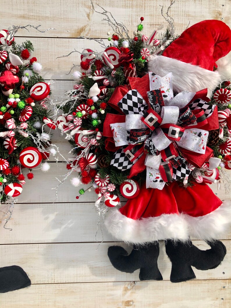 May include: A Christmas wreath decorated with candy canes, red and white swirl candies, and a Santa hat with black boots. The wreath is adorned with a black and white checkered bow.