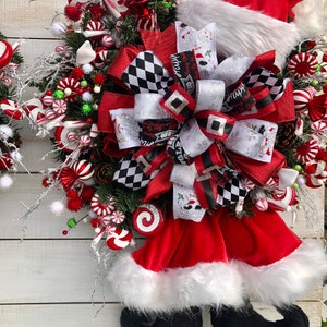 May include: A Christmas wreath with a red Santa hat, a red and black checkered bow, and candy cane decorations. The wreath is decorated with white pom-poms, green sprigs, and red and white candy canes.