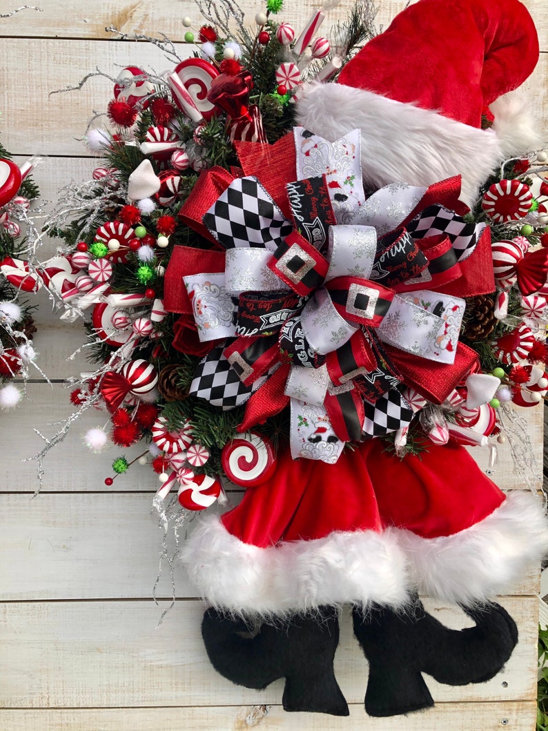 May include: A Christmas wreath with a red Santa hat, a red and black checkered bow, and candy cane decorations. The wreath is decorated with white pom-poms, green sprigs, and red and white candy canes.