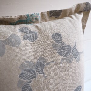 May include: A beige pillow with a floral pattern in shades of gray and teal. The pillow has a subtle texture and a simple design.