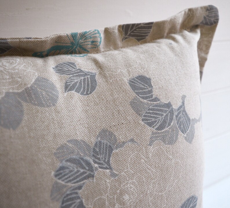 May include: A beige pillow with a floral pattern in shades of gray and teal. The pillow has a subtle texture and a simple design.
