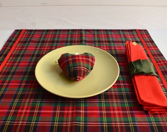 Pode incluir: Um tapete de mesa xadrez vermelho, verde e preto com um prato amarelo e um travesseiro em forma de coração xadrez vermelho, verde e preto no topo. Um guardanapo vermelho com uma fita verde está à direita do tapete de mesa.