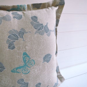 May include: A beige pillow with a blue and white floral pattern featuring butterflies. The pillow has a subtle linen texture.