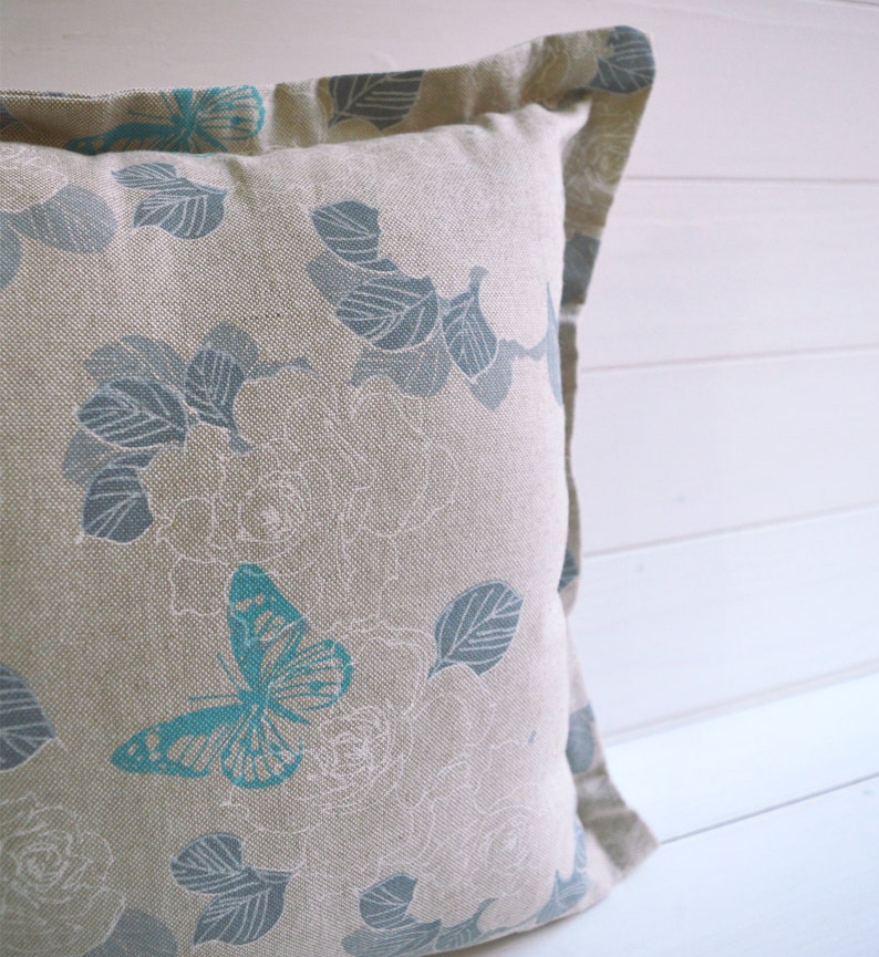 May include: A beige pillow with a blue and white floral pattern featuring butterflies. The pillow has a subtle linen texture.