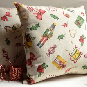 May include: Two beige throw pillows with a Christmas-themed pattern. The pattern includes a nutcracker, candy canes, presents, holly, and other festive elements.