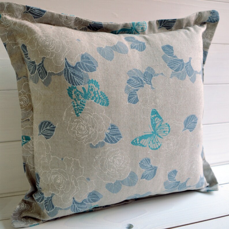 May include: A decorative throw pillow with a light gray linen fabric featuring a floral and butterfly pattern in shades of blue and teal.