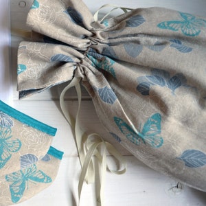 Peut inclure: Un sac en tissu beige à cordon avec un motif de papillon blanc et bleu. Le sac a un cordon blanc et est fait d'un tissu doux et texturé.