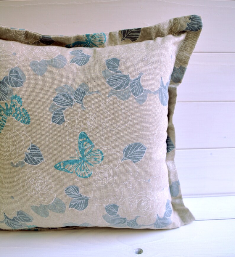 May include: A beige pillow with a floral pattern and blue butterflies. The pillow has a light brown border.