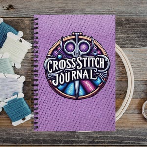 May include: A spiral-bound journal with a purple textured cover featuring a circular graphic of scissors and the text Cross Stitch Journal. The journal is surrounded by several spools of embroidery thread in various colors and a wooden hoop.