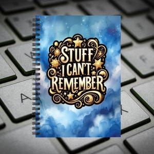 May include: A spiral-bound notebook with a blue watercolor-style background featuring a central gold-colored emblem with decorative swirls and stars. The text "STUFF I CAN'T REMEMBER" is printed in a bold, stylized font within the gold emblem.