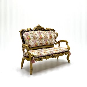 May include: A miniature sofa featuring a gold-colored frame with ornate carved details and a white fabric upholstery patterned with small colorful floral bouquets. The piece includes small pink tassels hanging from the front corners of the seat.