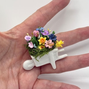 May include: Miniature white wheelbarrow filled with colorful flowers, including pink, yellow, blue, and purple blooms. The flowers sit atop green moss, creating a charming, handcrafted garden scene. The wheelbarrow is held in a hand.