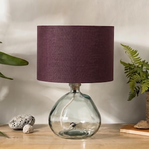 May include: A table lamp featuring a clear, rounded glass base with a visible white power cord inside and a cylindrical lampshade in a deep, solid plum purple fabric. The lamp has a simple, clean design with a smooth glass texture and fabric shade.