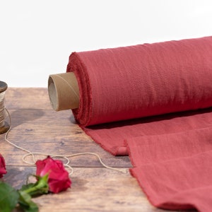 Könnte beinhalten: Eine Rolle roter Stoff mit rohem Rand, auf einer Holzoberfläche präsentiert. Eine Holzspule mit Faden und rote Rosen sind ebenfalls vorhanden. Der Stoff scheint Leinen oder eine ähnliche Naturfaser zu sein, für Näh- oder Bastelprojekte.