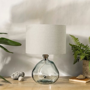 May include: A table lamp with a clear glass base and a white linen shade. The base is shaped like a bottle and has a textured surface. The lamp is sitting on a wooden table.
