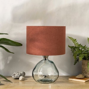 May include: A clear glass table lamp with a round base and a rust-coloured fabric lampshade. The lamp sits on a wooden surface, with plants and decorative stones in the background. The lamp's height is approximately 30 cm.