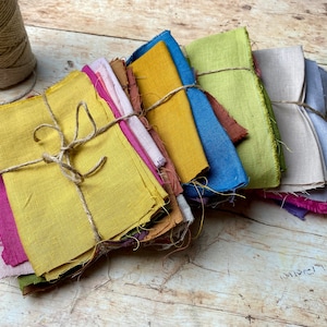 Op de afbeelding: Bundels gekleurde stoffen vierkanten in tinten geel, roze, blauw, groen en bruin, samengebonden met touw. De stof lijkt linnen of een vergelijkbare natuurlijke vezel te zijn. Een spoel touw ligt ernaast op een rustiek houten oppervlak.