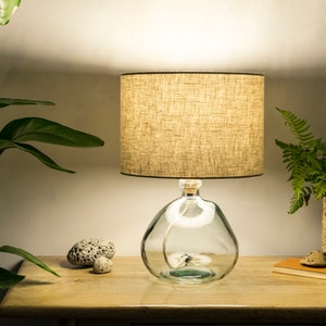 May include: A table lamp with a beige linen lampshade and a clear glass base. The base is shaped like a bottle and has a rope wrapped around it. The lamp is on a wooden table with a plant in a woven basket.