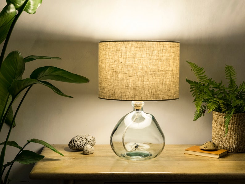 May include: A table lamp with a beige linen lampshade and a clear glass base. The base is shaped like a bottle and has a rope wrapped around it. The lamp is on a wooden table with a plant in a woven basket.