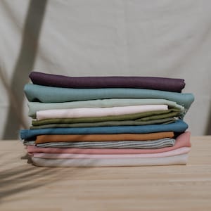 Puede incluir: Una pila de piezas de tela dobladas en varios colores, incluyendo morado oscuro, azul verdoso, verde claro, rosa p&aacute;lido, verde oliva, azul, amarillo mostaza, rayas grises y blancas, y rosa suave, dispuestas en un mont&oacute;n sobre madera.