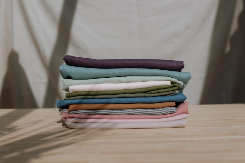 Puede incluir: Una pila de piezas de tela dobladas en varios colores, incluyendo morado oscuro, azul verdoso, verde claro, rosa p&aacute;lido, verde oliva, azul, amarillo mostaza, rayas grises y blancas, y rosa suave, dispuestas en un mont&oacute;n sobre madera.