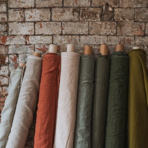 Puede incluir: Una colecci&oacute;n de siete rollos de tela verticales en varios colores, incluyendo gris claro, naranja &oacute;xido, crema, verde salvia, oliva oscuro y amarillo mostaza, cada uno enrollado alrededor de un tubo de cart&oacute;n y alineados uno al lado del otro.