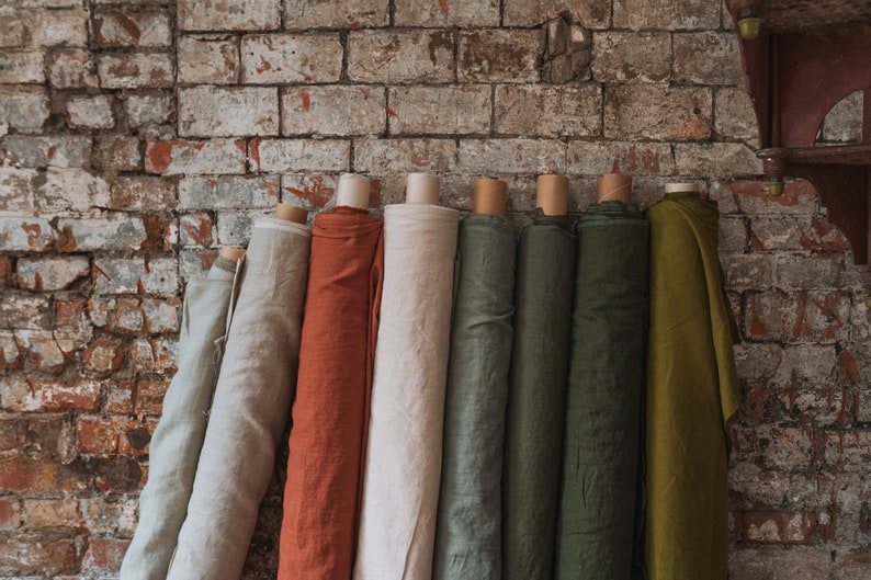 Puede incluir: Una colecci&oacute;n de siete rollos de tela verticales en varios colores, incluyendo gris claro, naranja &oacute;xido, crema, verde salvia, oliva oscuro y amarillo mostaza, cada uno enrollado alrededor de un tubo de cart&oacute;n y alineados uno al lado del otro.