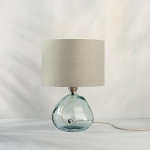 May include: A table lamp with a light grey linen shade and a clear glass base with a textured surface.