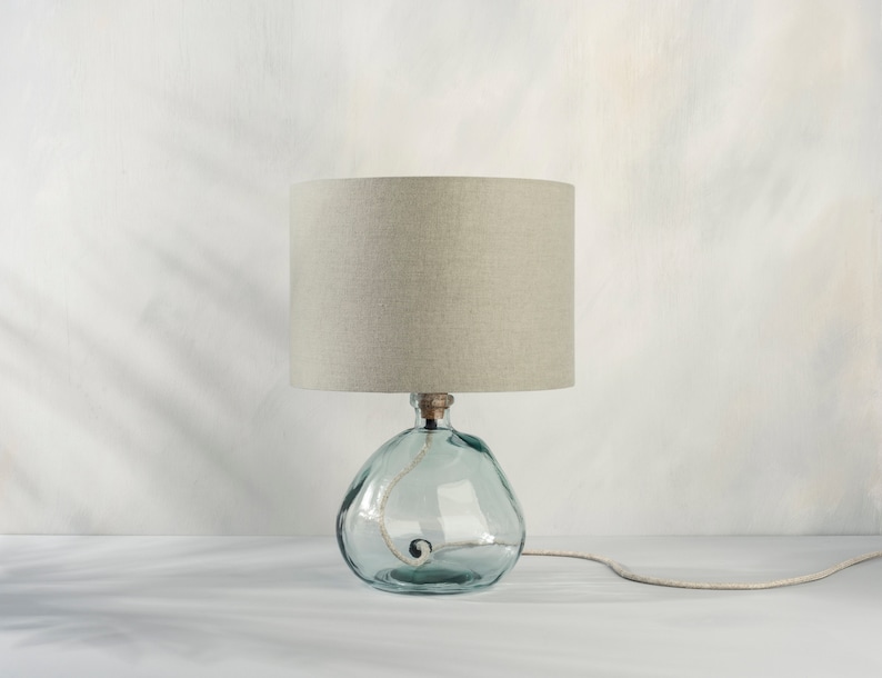 May include: A table lamp with a light grey linen shade and a clear glass base with a textured surface.