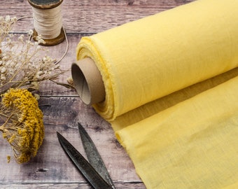 May include: A roll of yellow fabric on a wooden surface, with scissors, dried flowers, and a thread spool. The fabric is a light, solid yellow, possibly linen. The scissors are open, and the scene suggests a craft or sewing project.