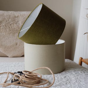 May include: Two fabric lampshades, one olive green and one beige, are stacked on a bed. A coiled brown electrical cord with a plug is in the foreground. A black jug with dried flowers sits on a small wooden table.