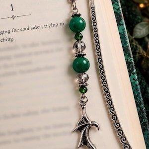 May include: A silver metal hook bookmark featuring a decorative scroll pattern. A dangling charm chain includes two large green beads, two smaller faceted dark gray beads, two tiny green crystal beads, and a silver dragon wing charm.