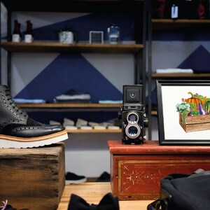 May include: A framed print of a watercolor painting of a wooden crate filled with various vegetables, including a pumpkin, eggplant, bell peppers, and broccoli. The print is displayed on a shelf in a retail store.