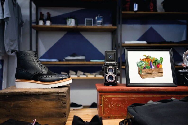 May include: A framed print of a watercolor painting of a wooden crate filled with various vegetables, including a pumpkin, eggplant, bell peppers, and broccoli. The print is displayed on a shelf in a retail store.